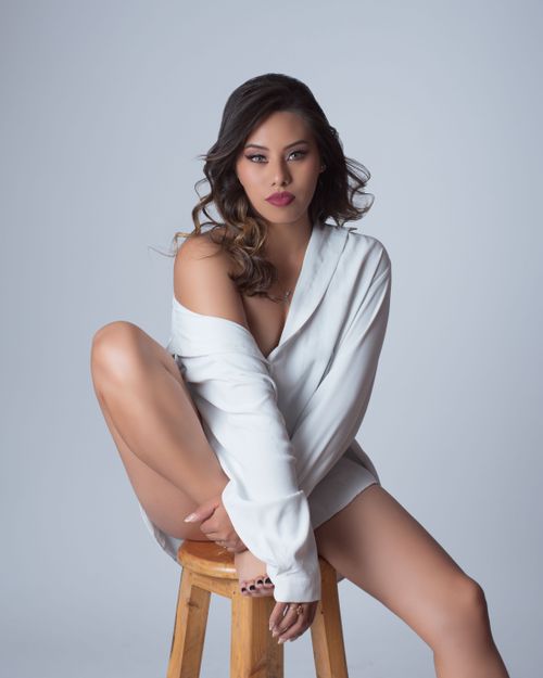 Free Elegant portrait of a woman seated on a stool wearing a white shirt showcasing a modern and confident style. Stock Photo