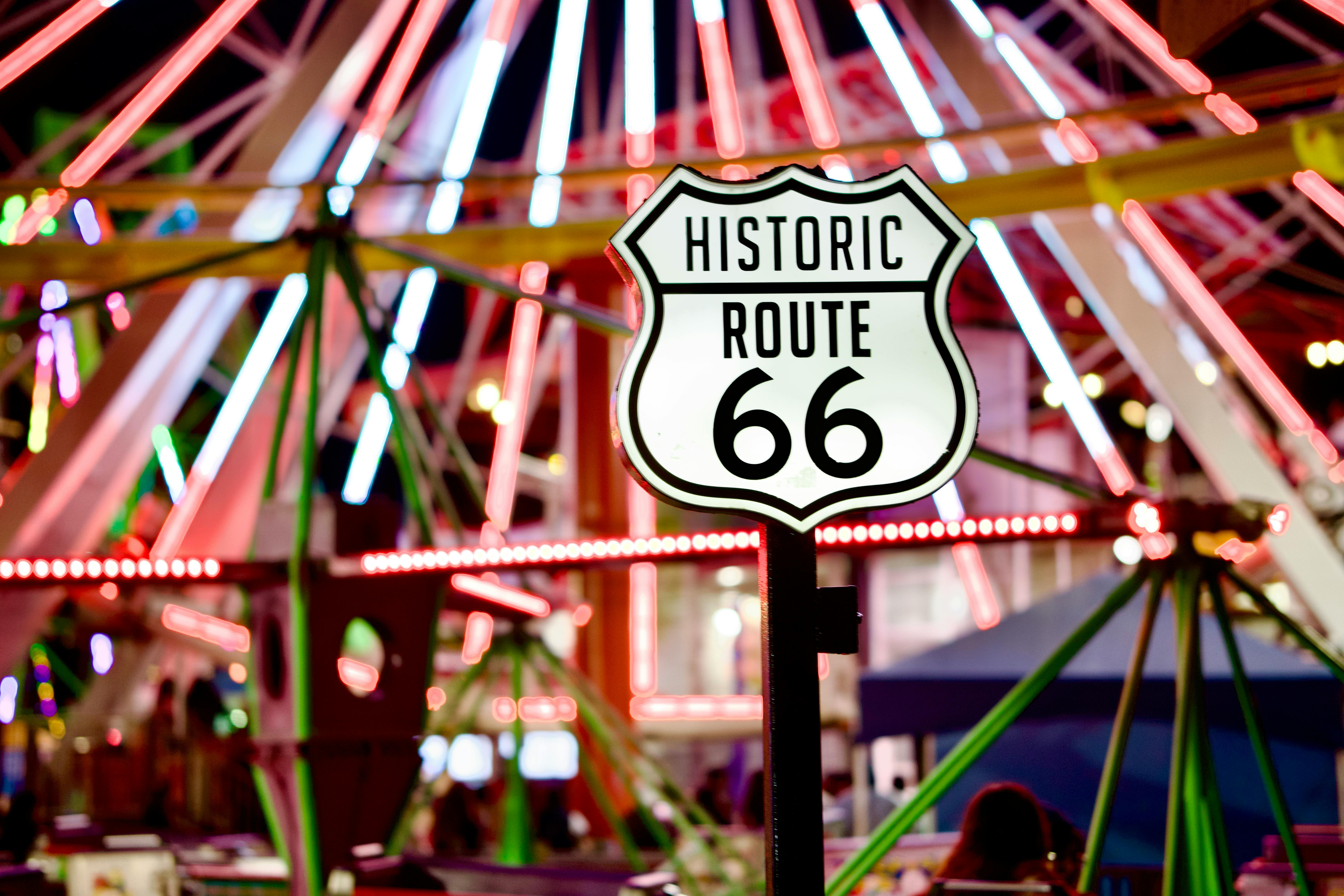 Historic Route 66 sign illuminated at a colorful night carnival with ferris wheel lights.
