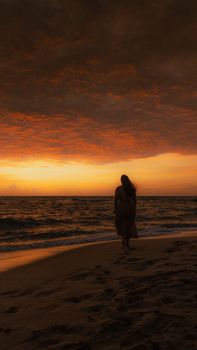 A woman walks along a beach at sunset, creating a dramatic silhouette against the golden sky.