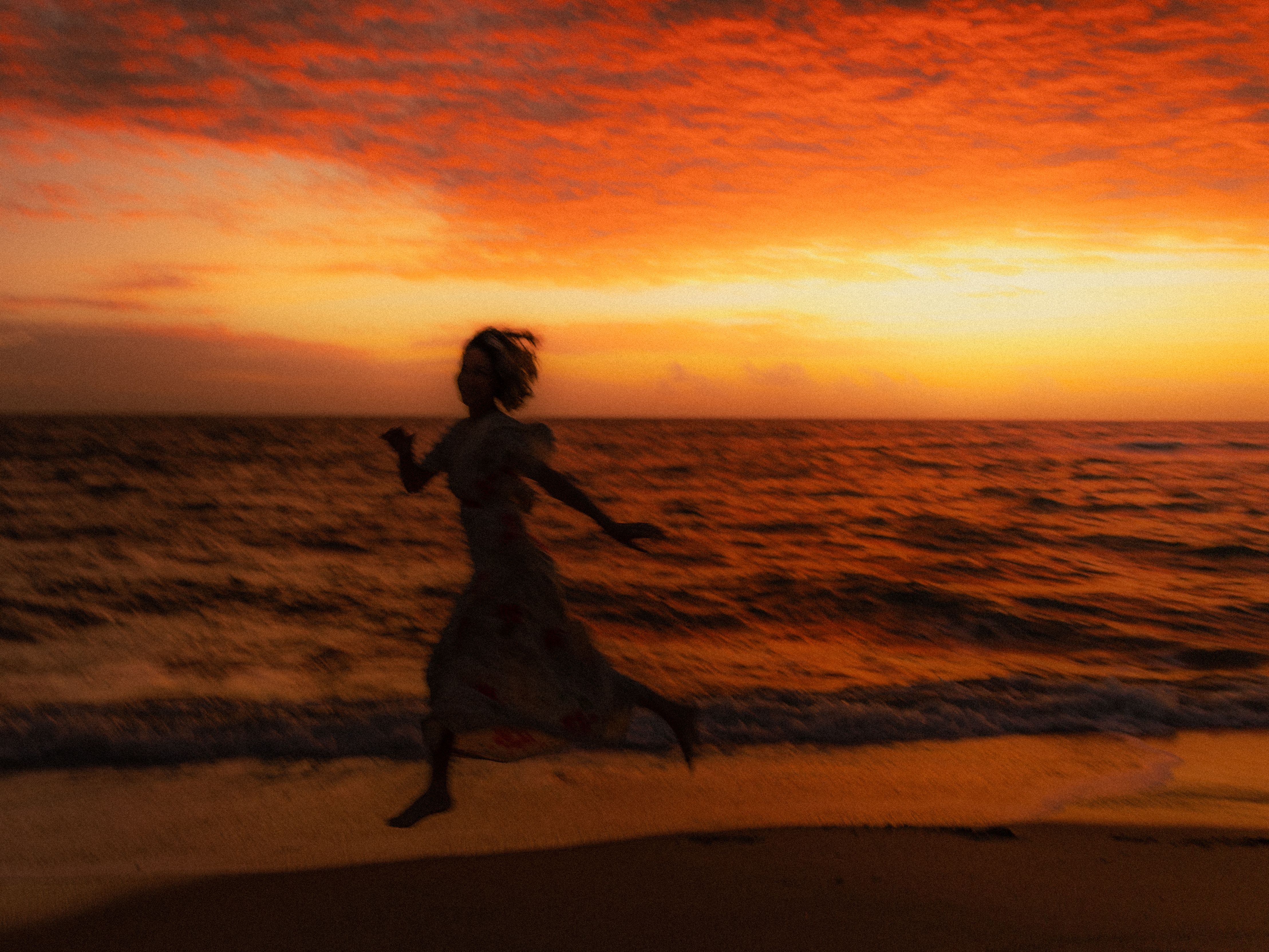 Gratis Una donna si staglia contro un tramonto vibrante, mentre corre lungo la spiaggia sotto un cielo rosso intenso. Foto a disposizione