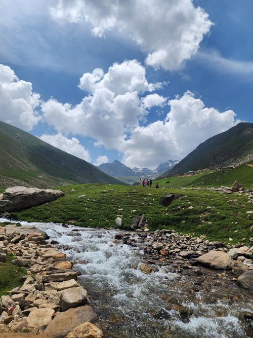 Free A picturesque mountain stream flows through a vibrant green valley with stunning cloud formations overhead. Stock Photo