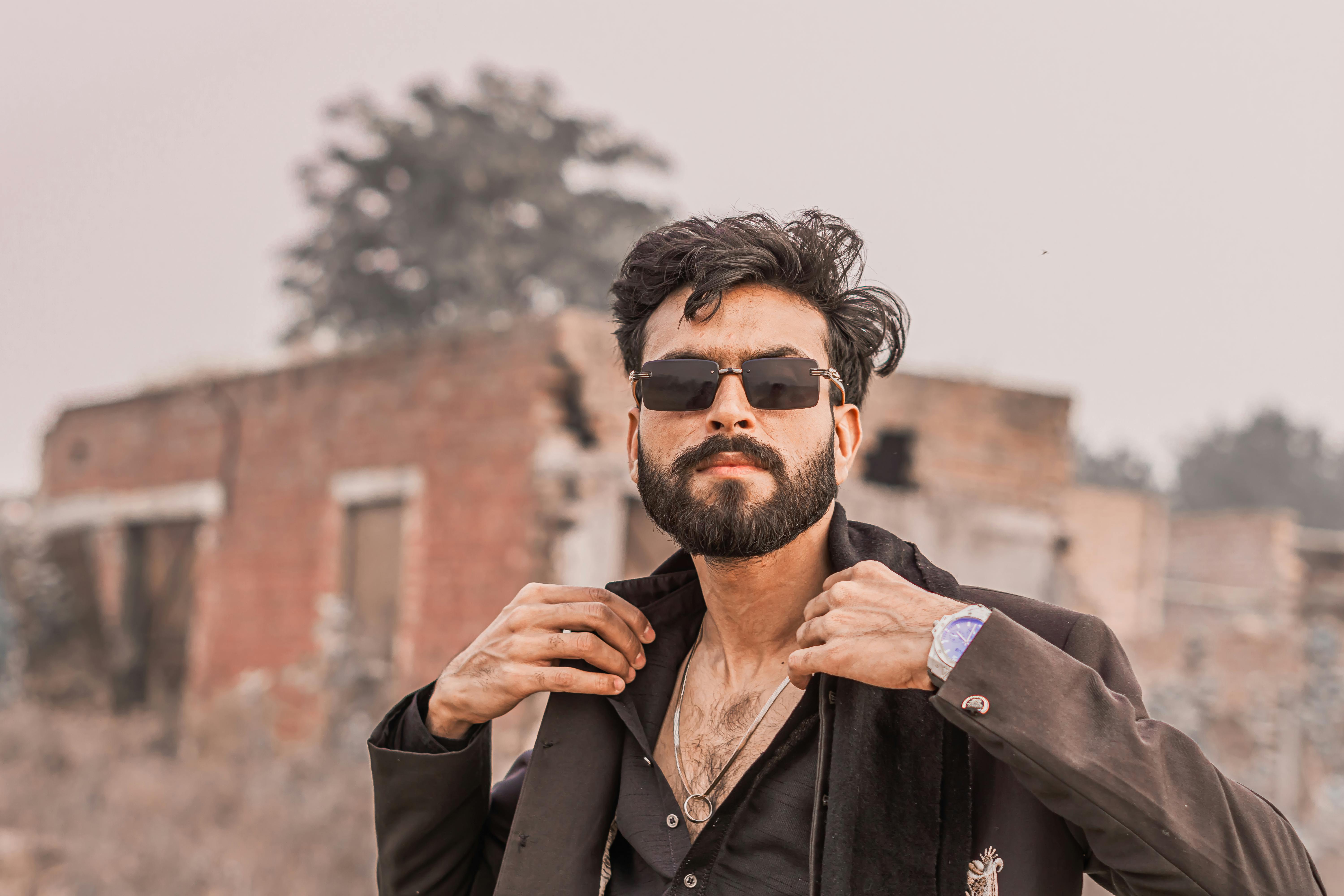 Confident man in sunglasses adjusting jacket outdoors near rustic buildings.