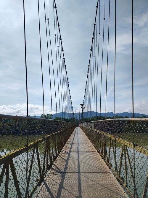 Free A stunning view of a suspension bridge spanning over a serene river with mountains in the background. Stock Photo