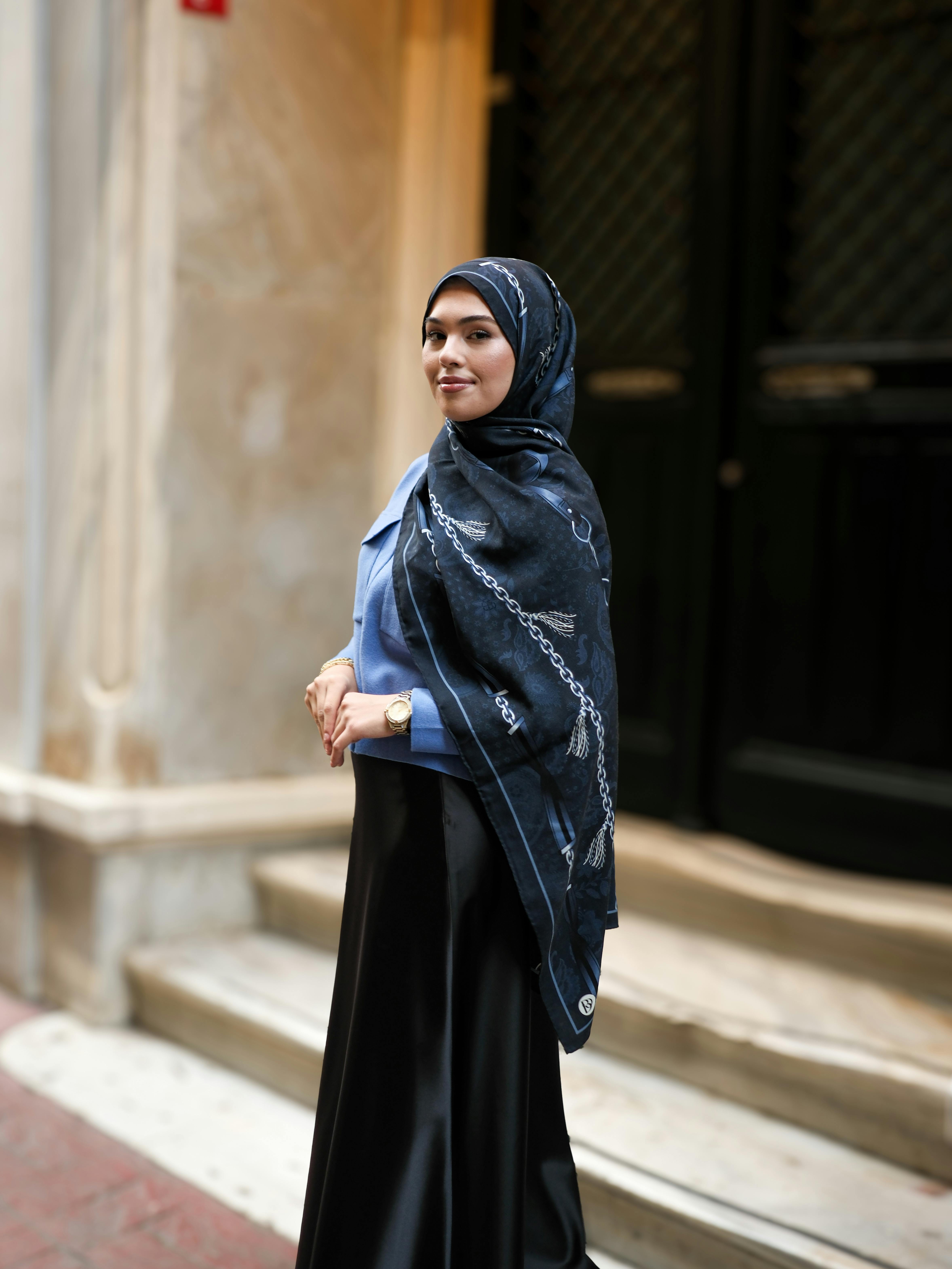 A stylish woman wearing a hijab stands confidently on a city street, portraying grace and modern fashion.