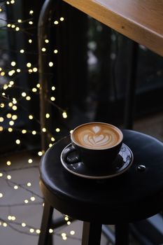 A cup of coffee with latte art on a stool, surrounded by warm string lights.