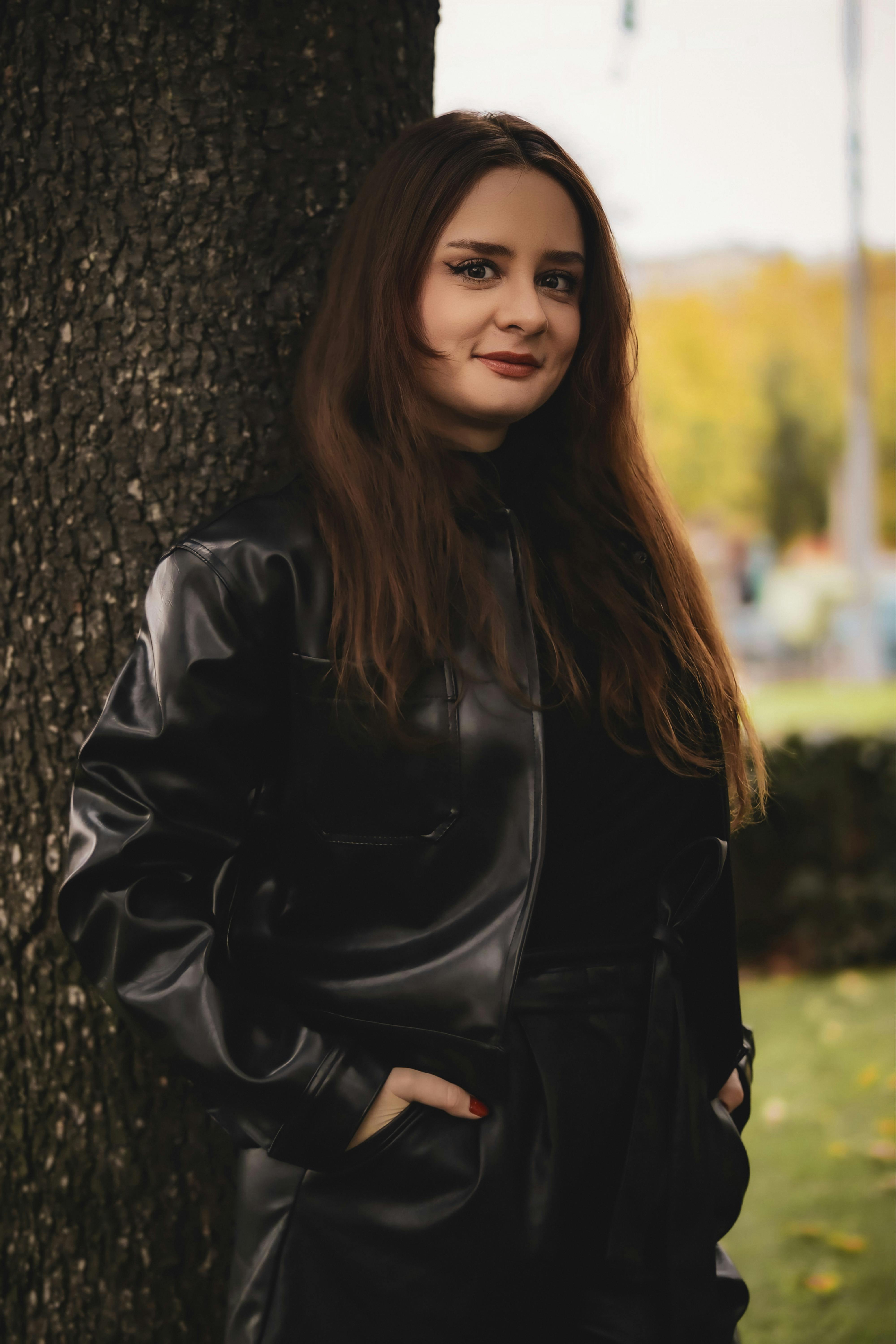 A woman in a black leather jacket poses by a tree in a park during fall.
