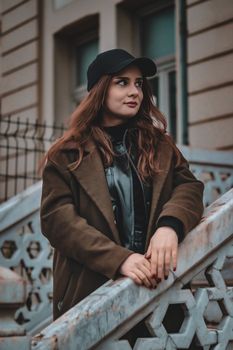 Young woman in a brown coat and black cap poses on marble stairs outdoors, showcasing modern fashion.