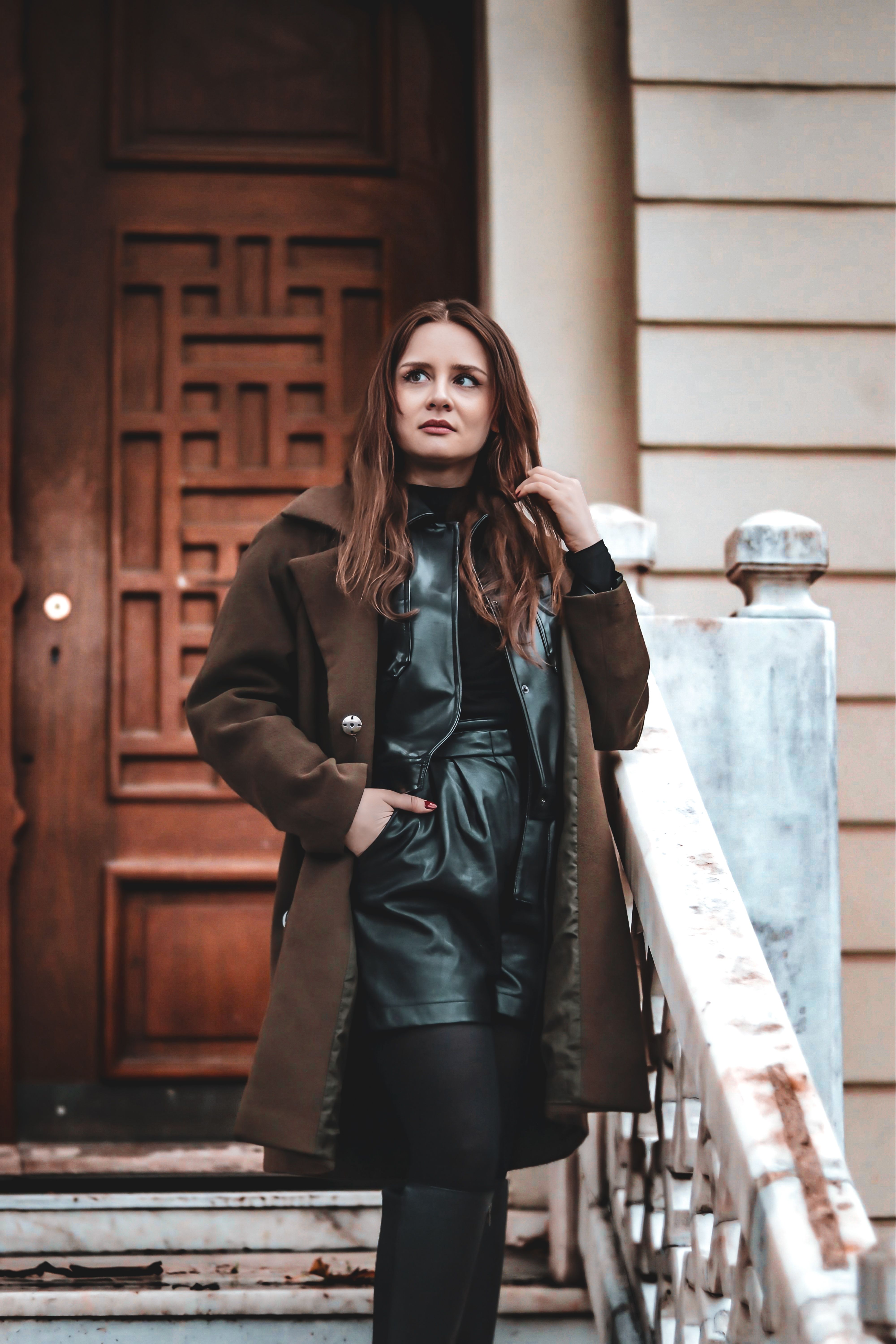 A fashionable woman in a brown coat poses on outdoor stairs, exuding confidence and style.