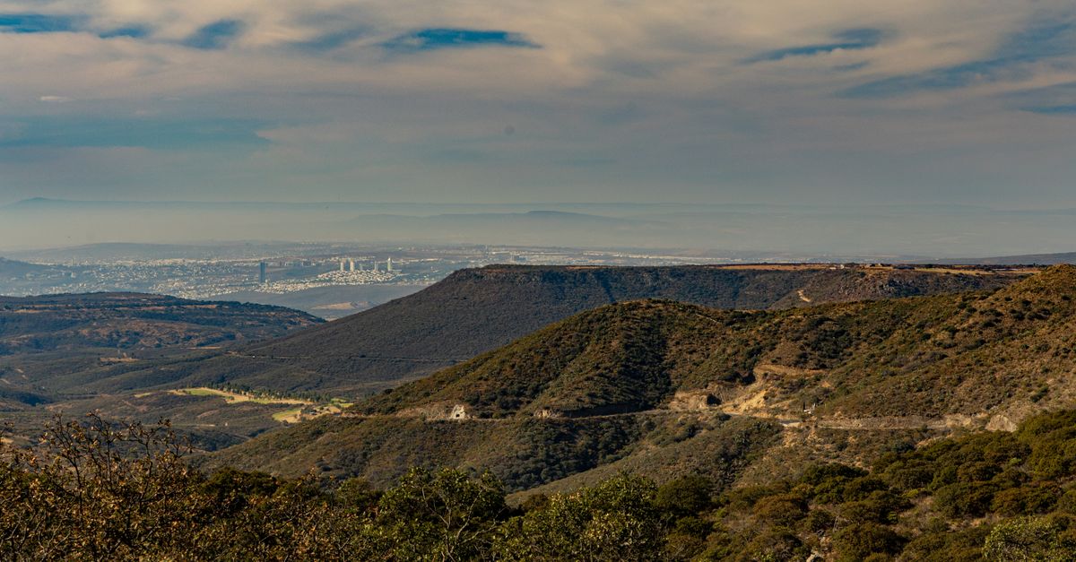 Aerial view of mountainous terrain with a distant cityscape under a cloudy sky.