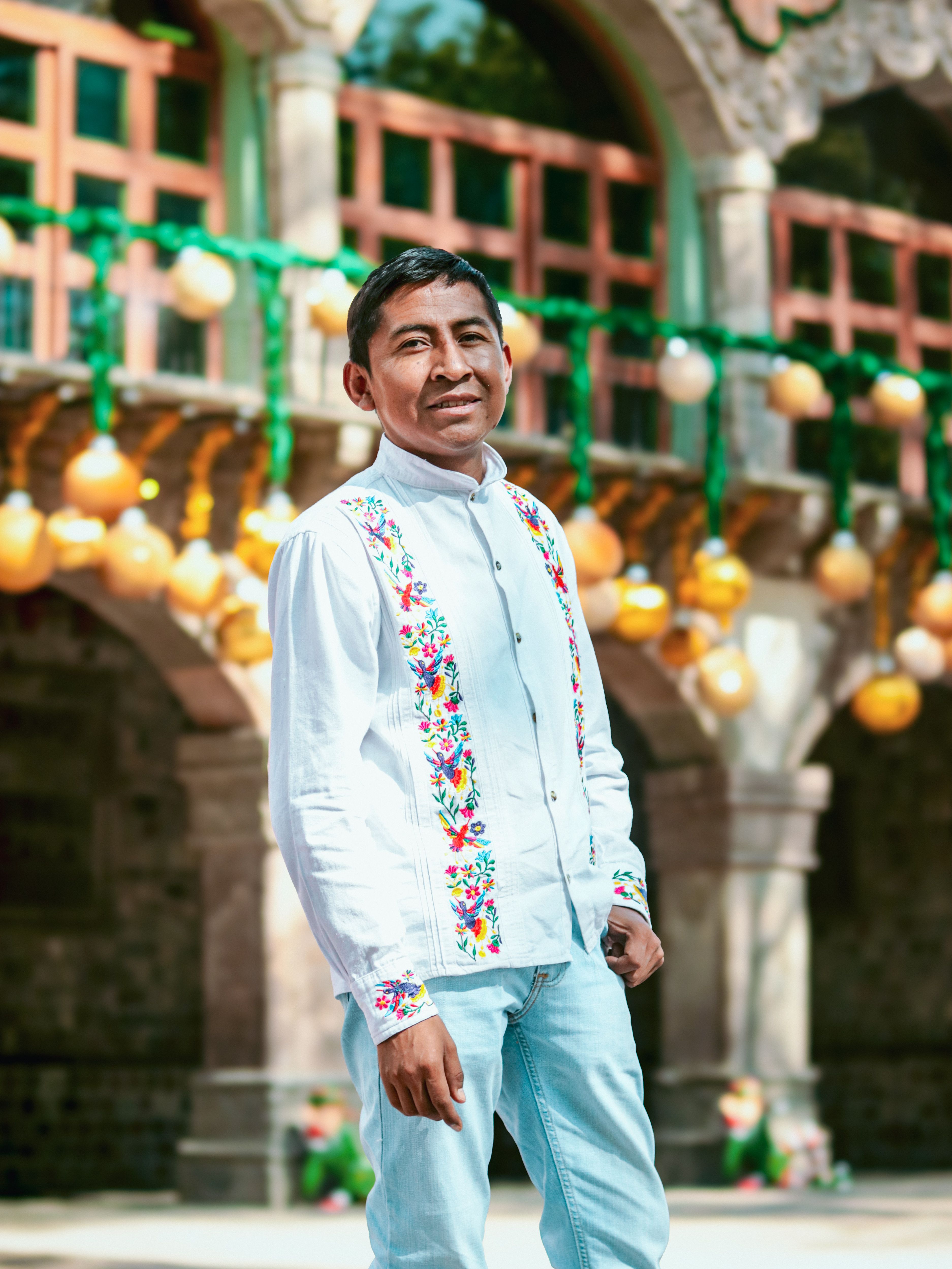 Man in traditional embroidered shirt poses outdoors with festive decor. Bright, cultural atmosphere captured.