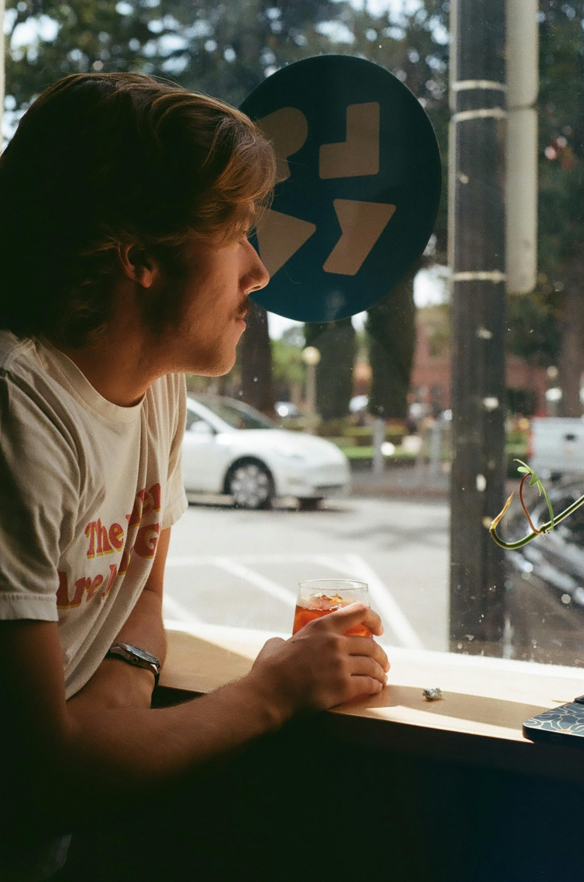 Young man enjoying a drink at a cafe window overlooking a street.