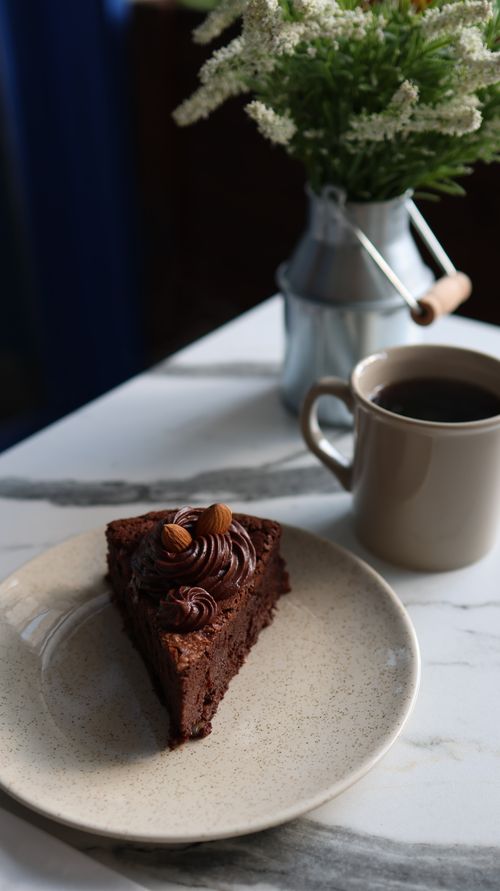 Free A slice of rich chocolate tart with almonds, paired with a cup of coffee and floral decor on a marble table. Stock Photo