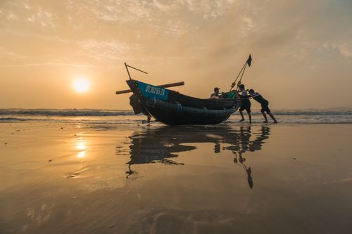 Free Fishermen pushing a traditional boat ashore during a golden sunset, with the sea reflecting the tranquil scene. Stock Photo