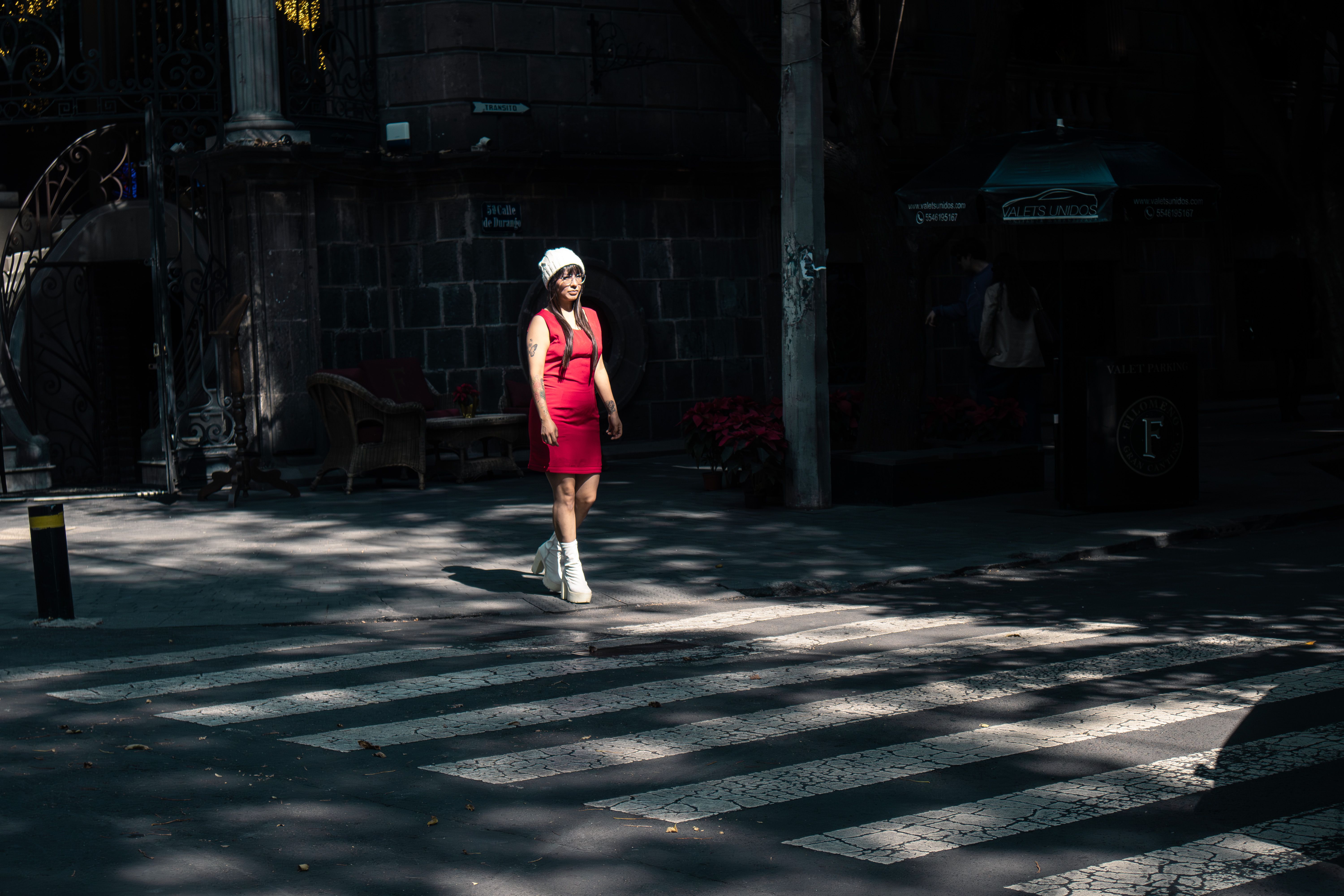 A woman in a red dress and white hat crosses a city street in bright daylight.