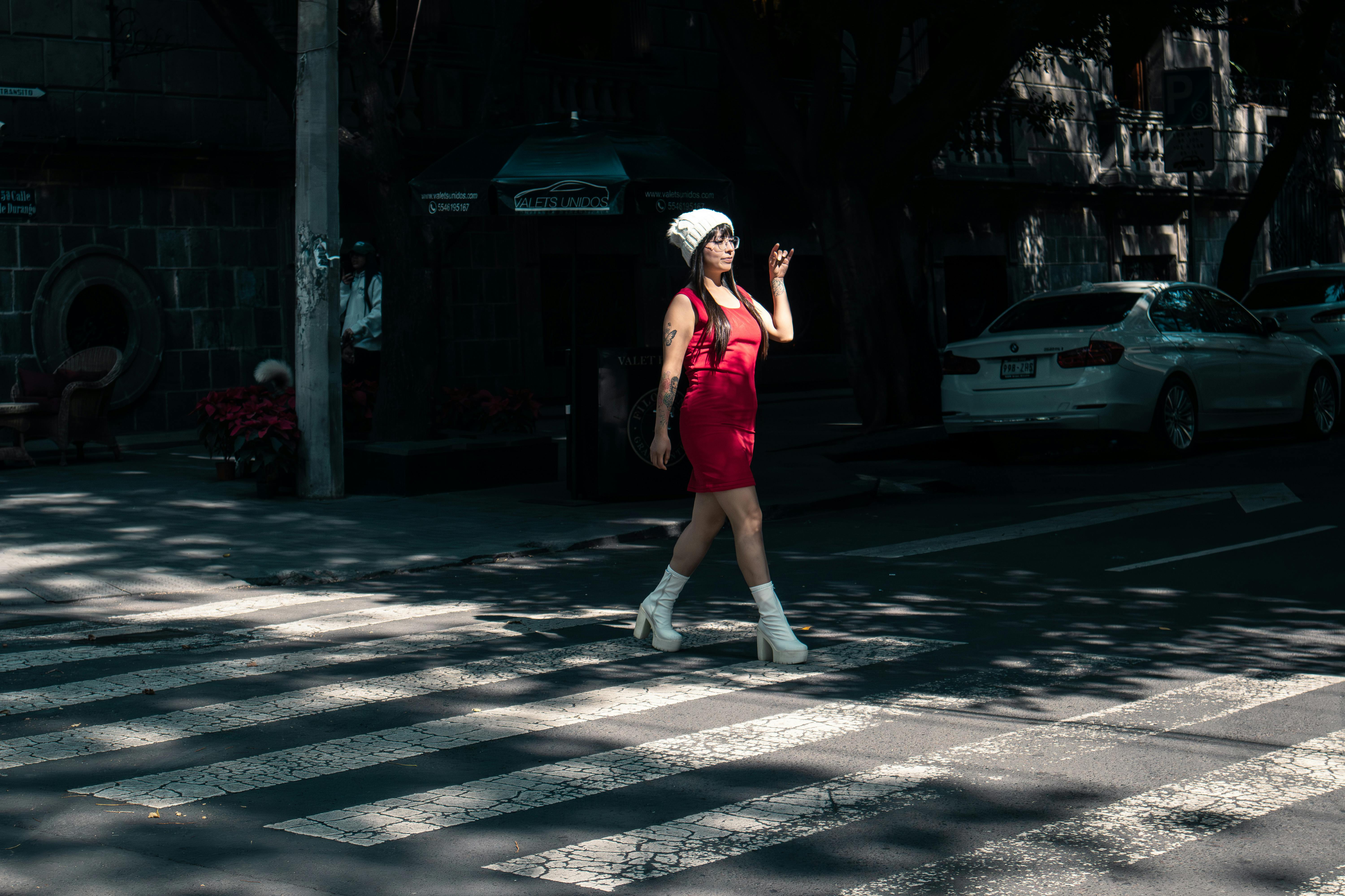 Fashionable woman in red dress with white boots crossing a city street in daylight.