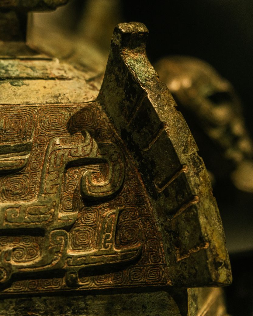 The rim of an ancient cast bronze vessel, its raised geometric ornament mottled green and gold where patina has formed over the metal.