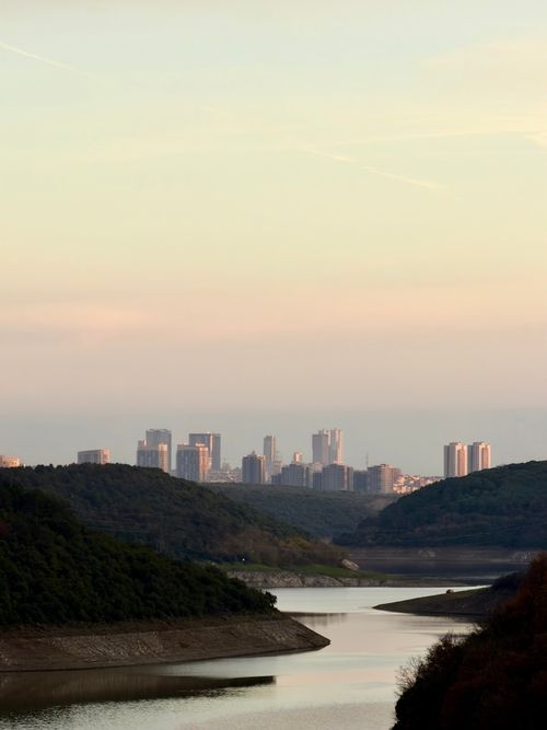 Free Serene river view with distant city skyline at dusk, surrounded by lush green hills. Stock Photo