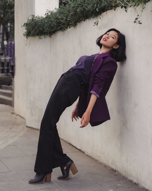 Free Woman in Purple Long Sleeve Shirt and Black Pants Leaning on White Wall Stock Photo
