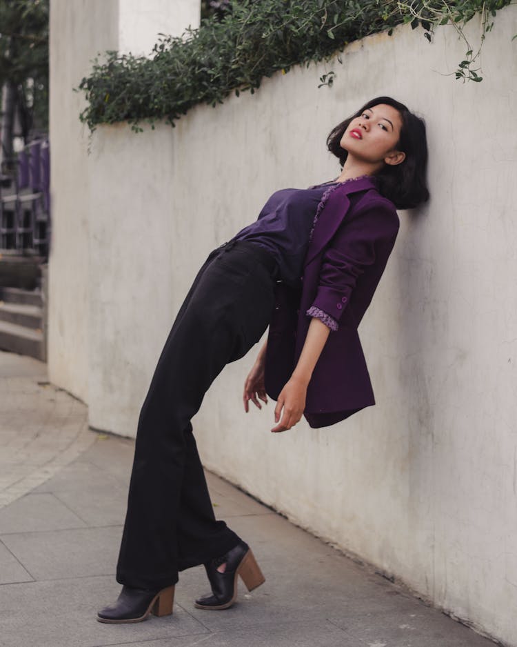 Woman In Purple Long Sleeve Shirt And Black Pants Leaning On White Wall