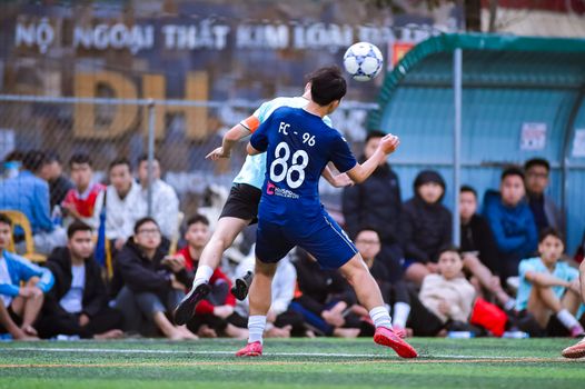 Dynamic action shot of amateur football game in Hanoi, Vietnam with two players engaged and fans watching.