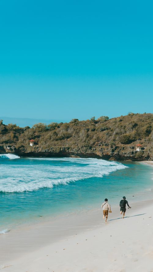 Free A peaceful beach scene in Indonesia with two people walking along the shore, under a clear sky. Stock Photo