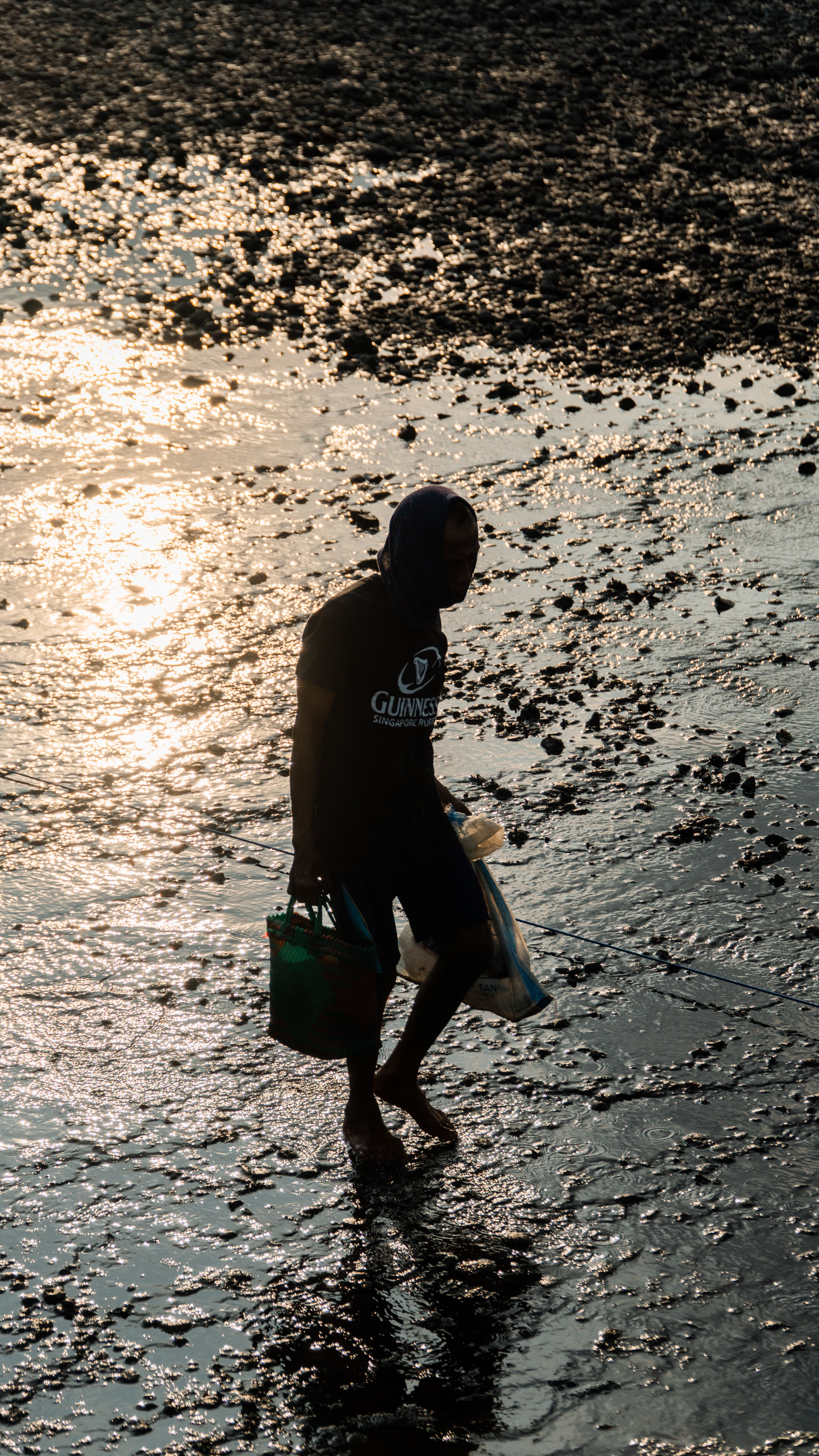 A man silhouetted against the setting sun walks across a wet, rocky shore in Indonesia.