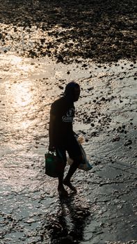 A man silhouetted against the setting sun walks across a wet, rocky shore in Indonesia.