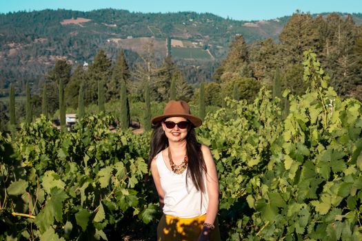 A woman in a hat and sunglasses smiles amidst lush vineyard with scenic mountain backdrop.