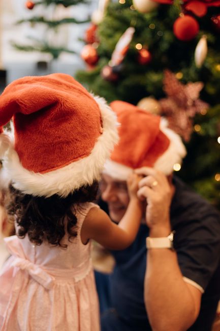 A child and adult share festive moments by a decorated Christmas tree wearing Santa hats.