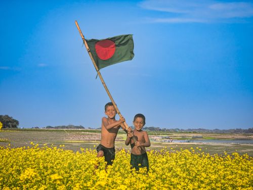 Free Two boys joyfully wave the Bangladesh flag in a vibrant mustard field under a clear blue sky. Stock Photo