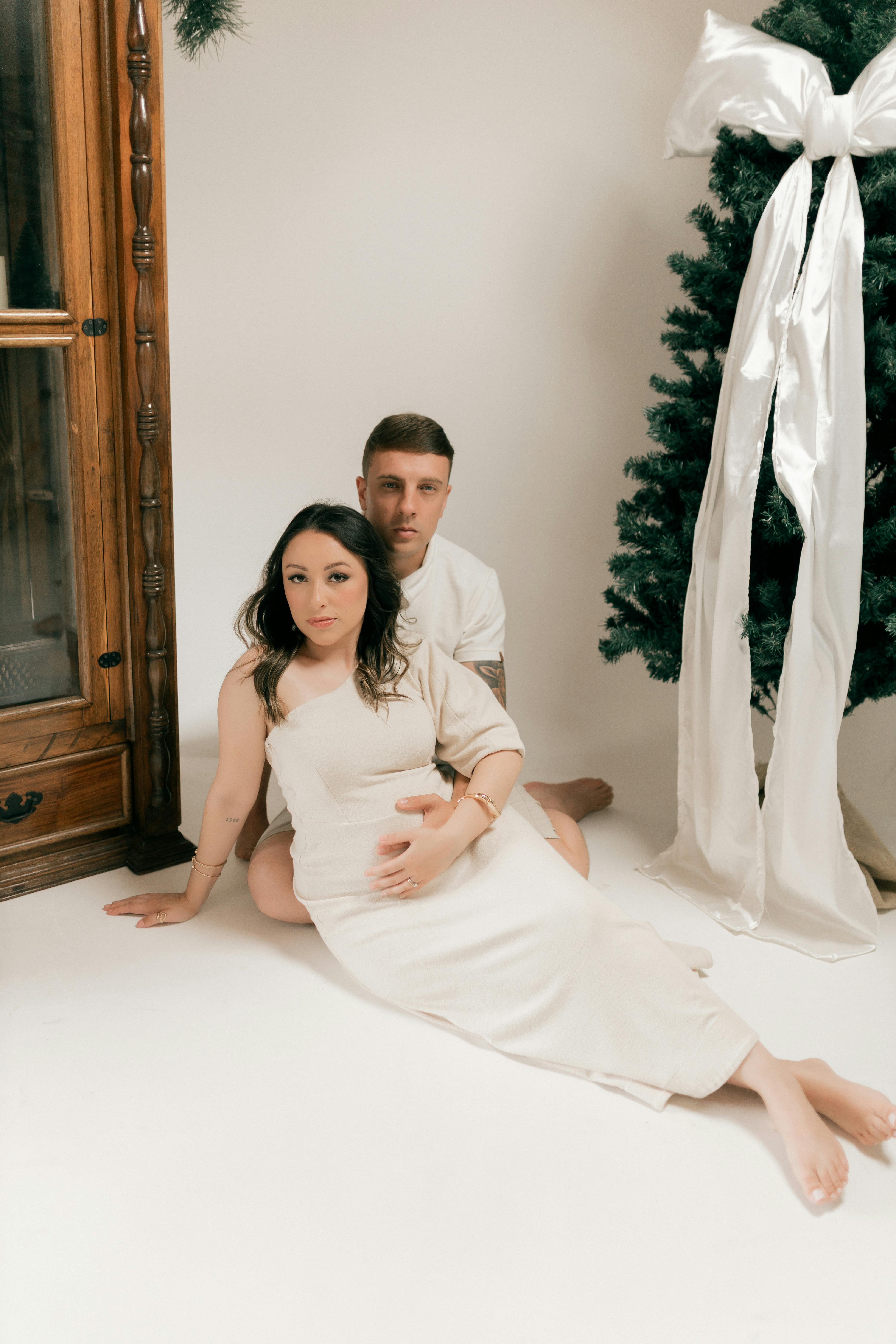 Expectant couple sitting by a decorated Christmas tree, celebrating togetherness and upcoming family joy.