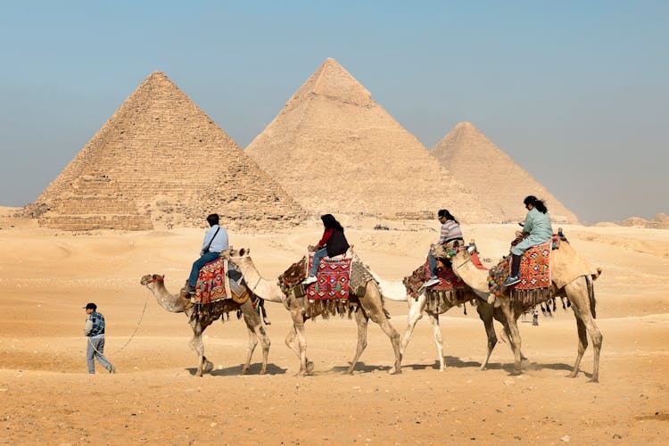 Four People Riding On Camels Across The Pyramids