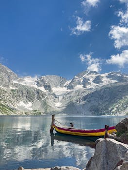 A vibrant boat floats on a serene mountain lake with snowy peaks in the background.