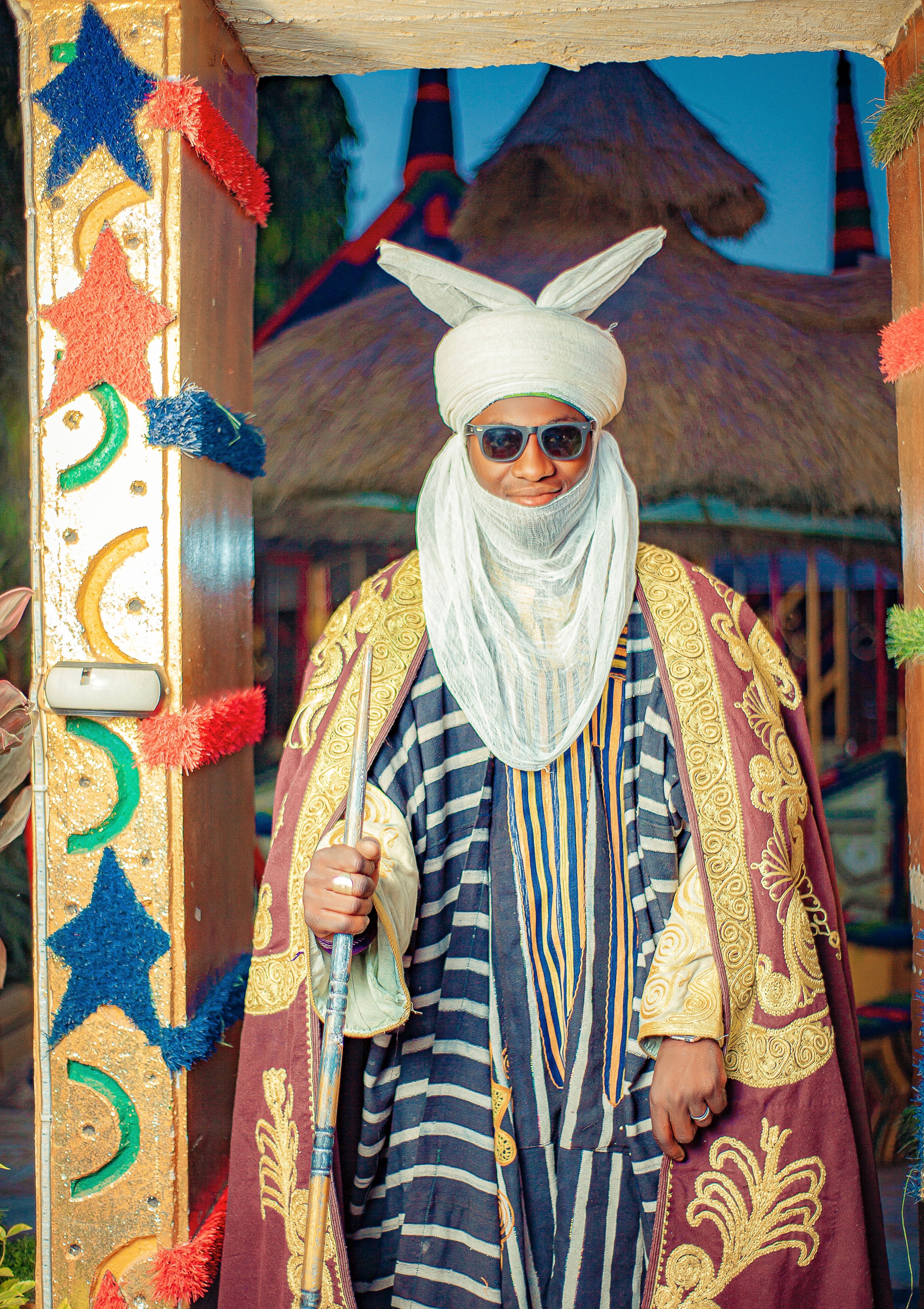 Man in ceremonial Nigerian attire stands by colorful traditional building.