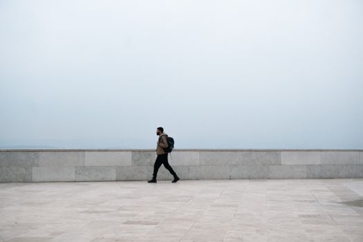 A man strolls along a minimalist outdoor stone walkway under an overcast sky.