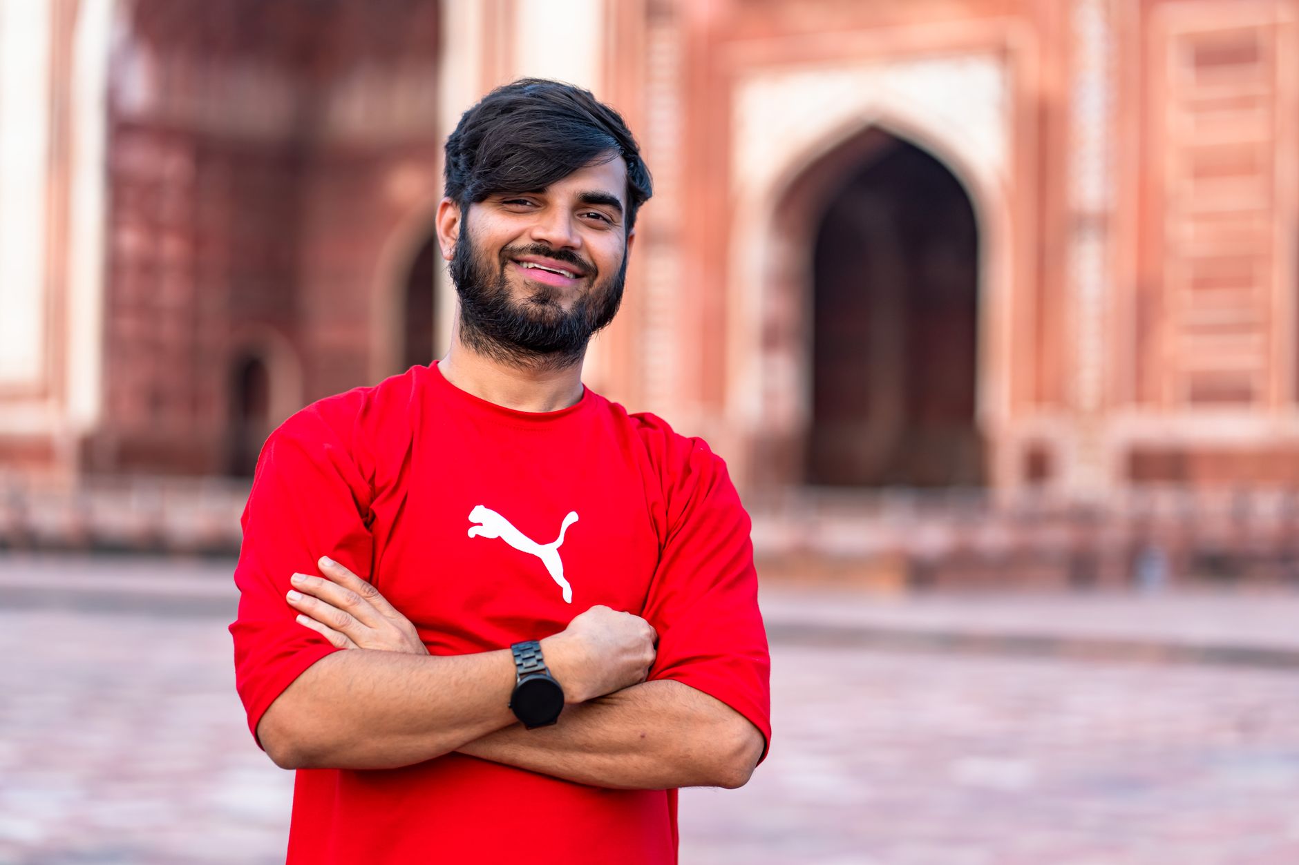 Smiling man in red shirt stands confidently before historic building with arches.