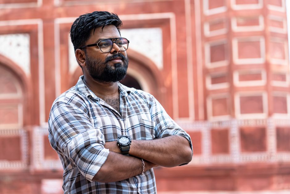 Man with glasses confidently standing in front of a historic building in a plaid shirt.
