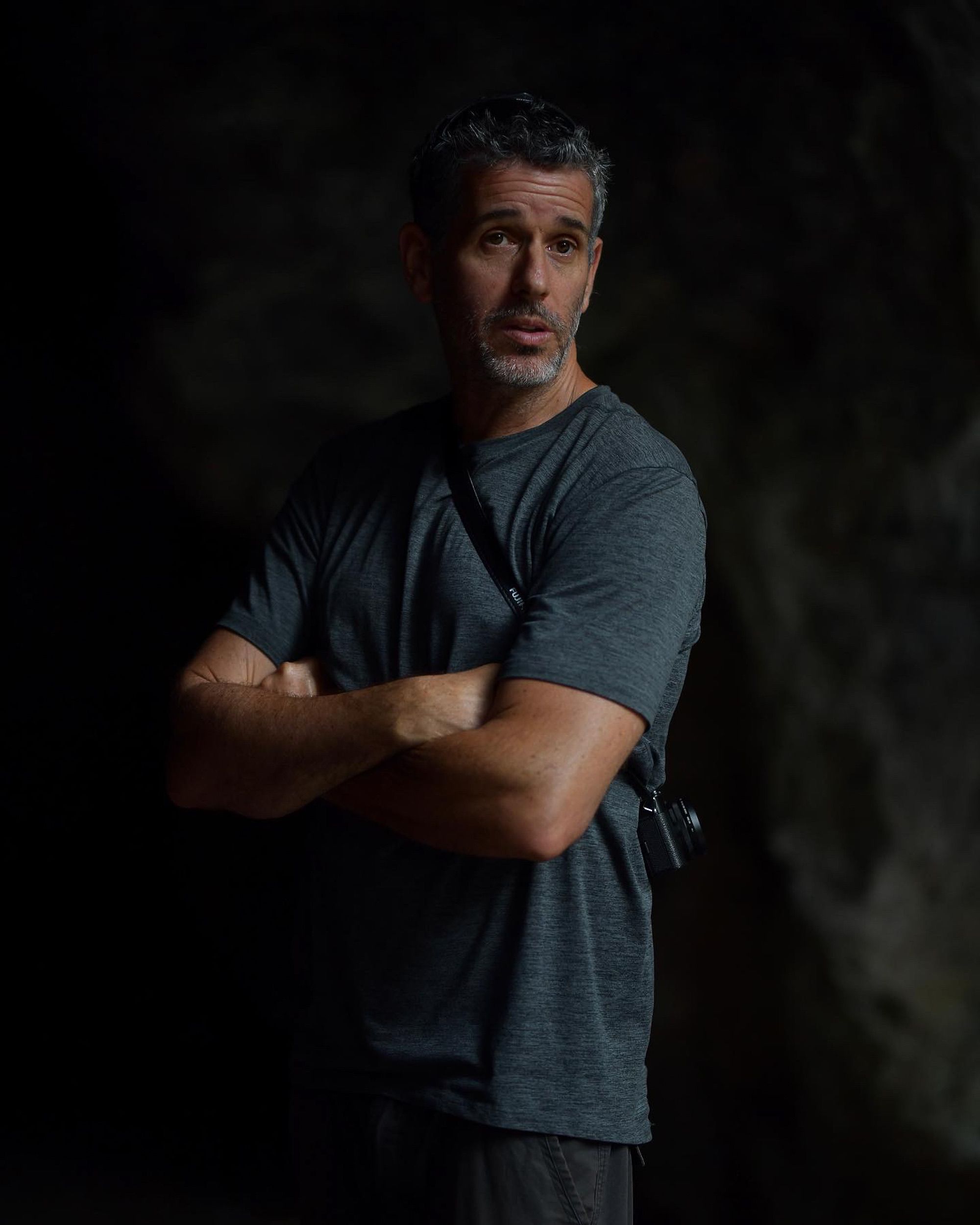 Man with crossed arms stands pensively in a dark cave setting. Low light emphasizes mood.