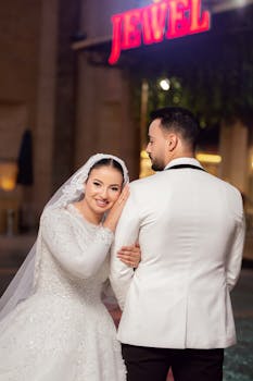 Bride and groom in elegant attire posing under glowing sign at outdoor night wedding.