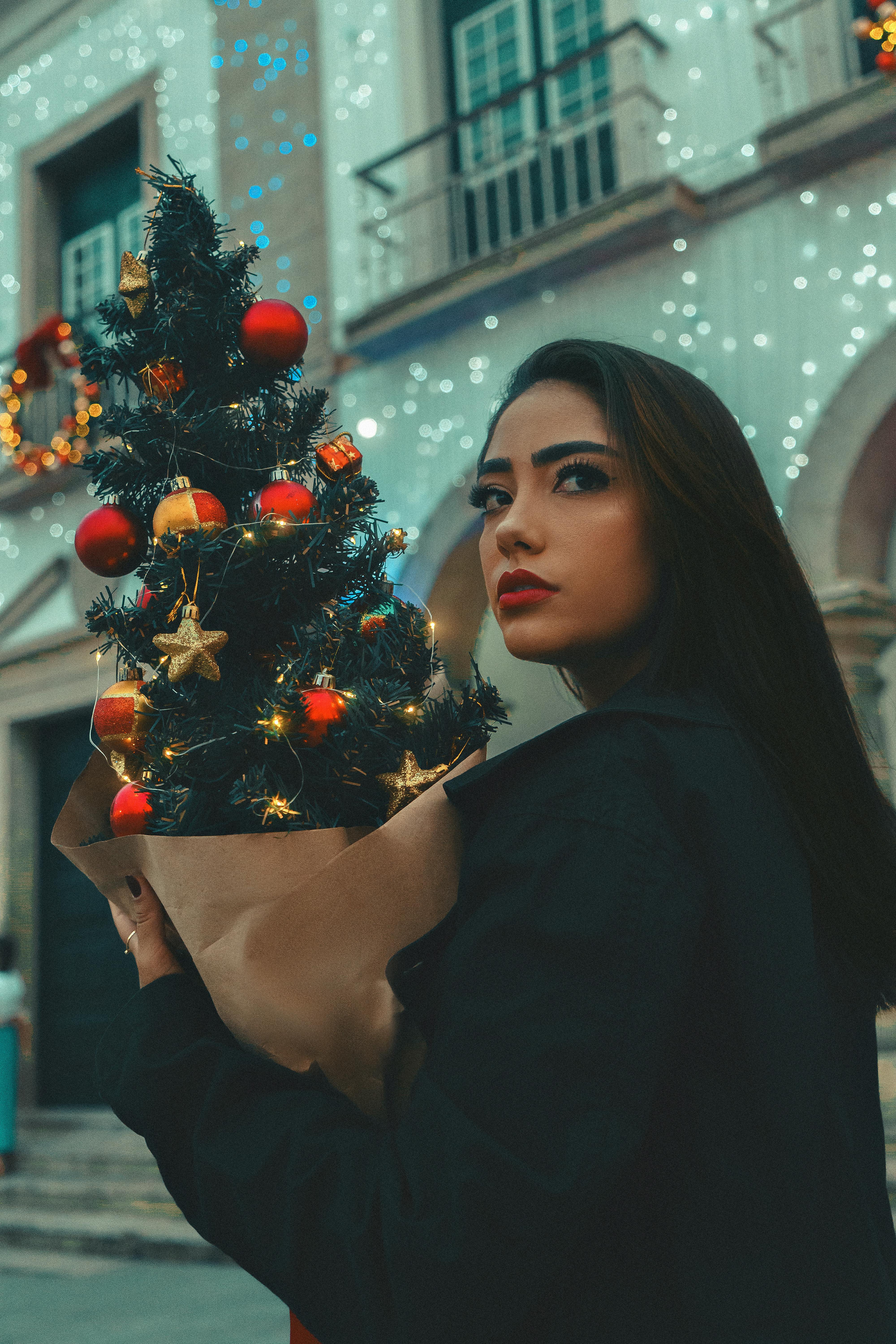 A woman holding a decorated mini Christmas tree, standing outdoors with festive lights in the background.