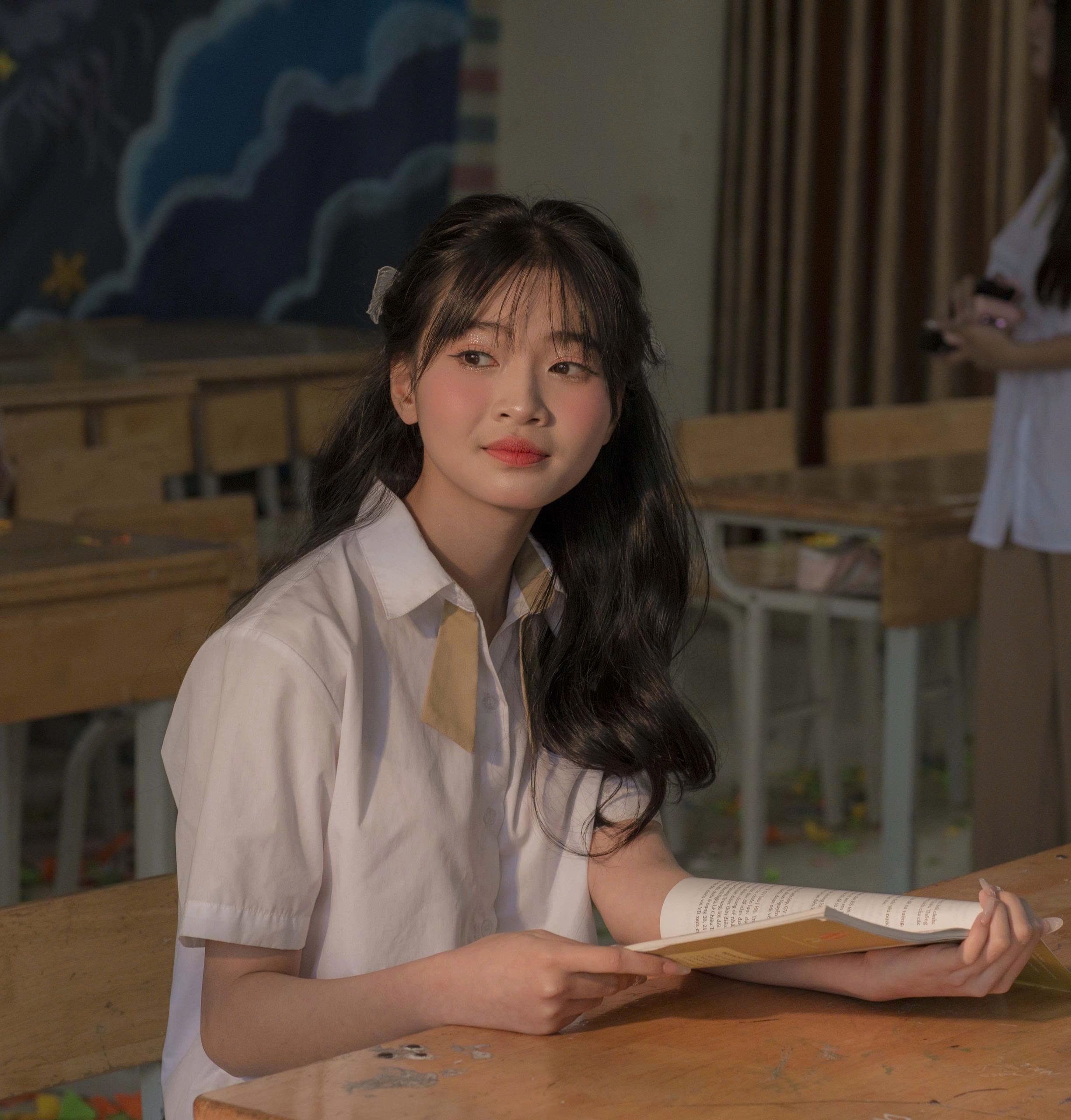 A young student sits thoughtfully in a dimly lit classroom holding a book.