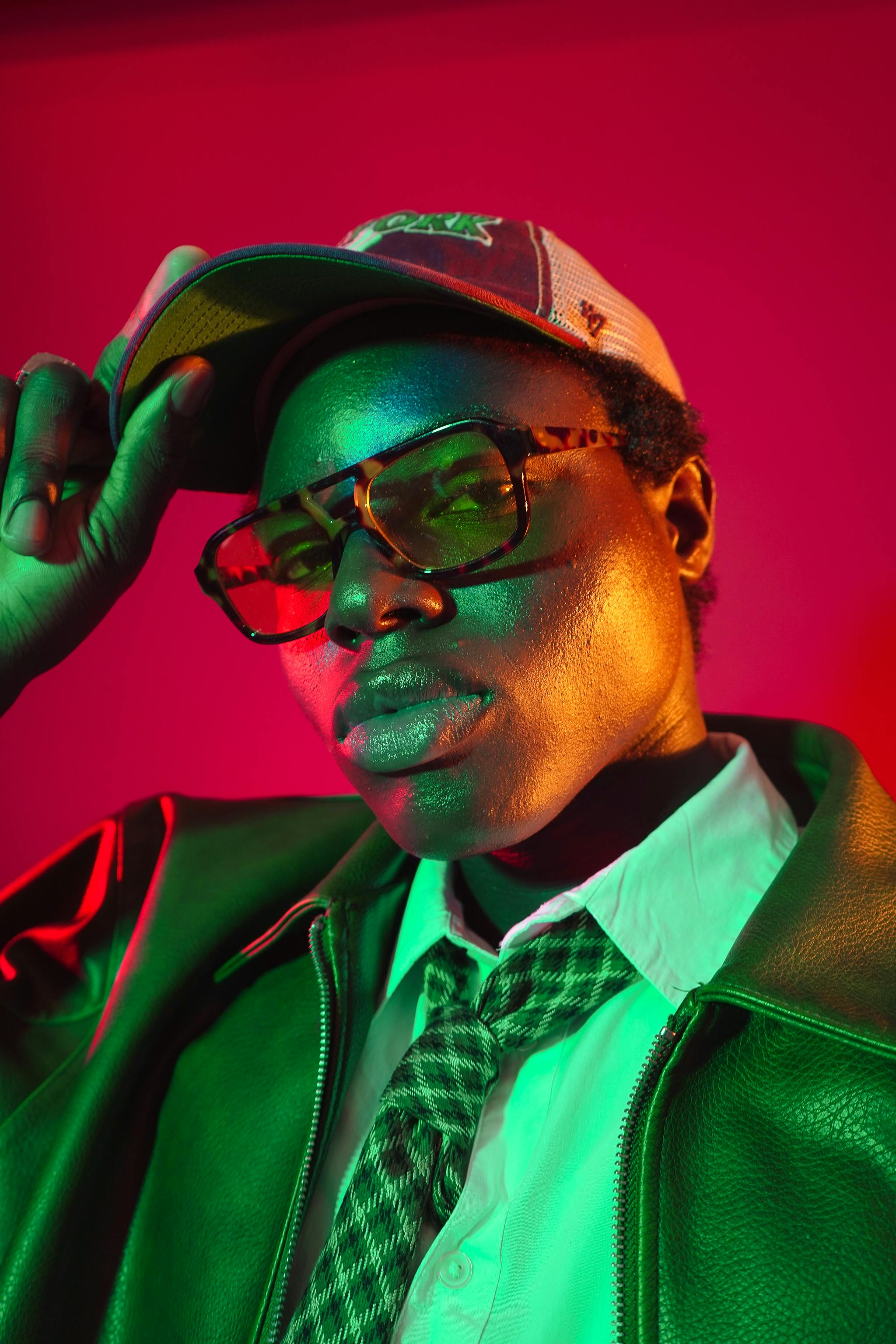Editorial fashion portrait of a model in colorful studio lighting wearing a cap and sunglasses.