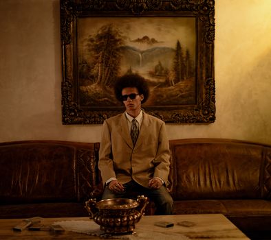 Man in a tan suit and sunglasses sits on an elegant sofa in a vintage living room.