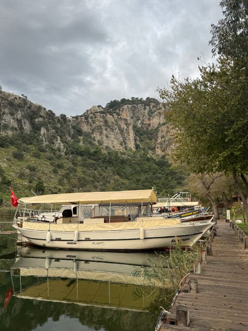 Free A tranquil riverside scene with a boat docked by lush mountains, under a cloudy sky. Stock Photo