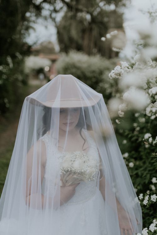 Free Bride in white dress holding bouquet under a veil in serene garden setting. Stock Photo