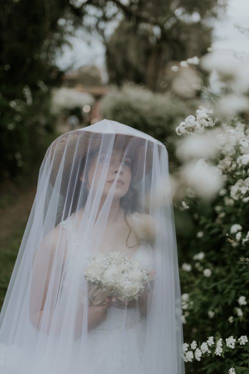 Free Bride in wedding dress and veil holding bouquet amidst lush greenery and blooming flowers, creating a soft and romantic scene. Stock Photo