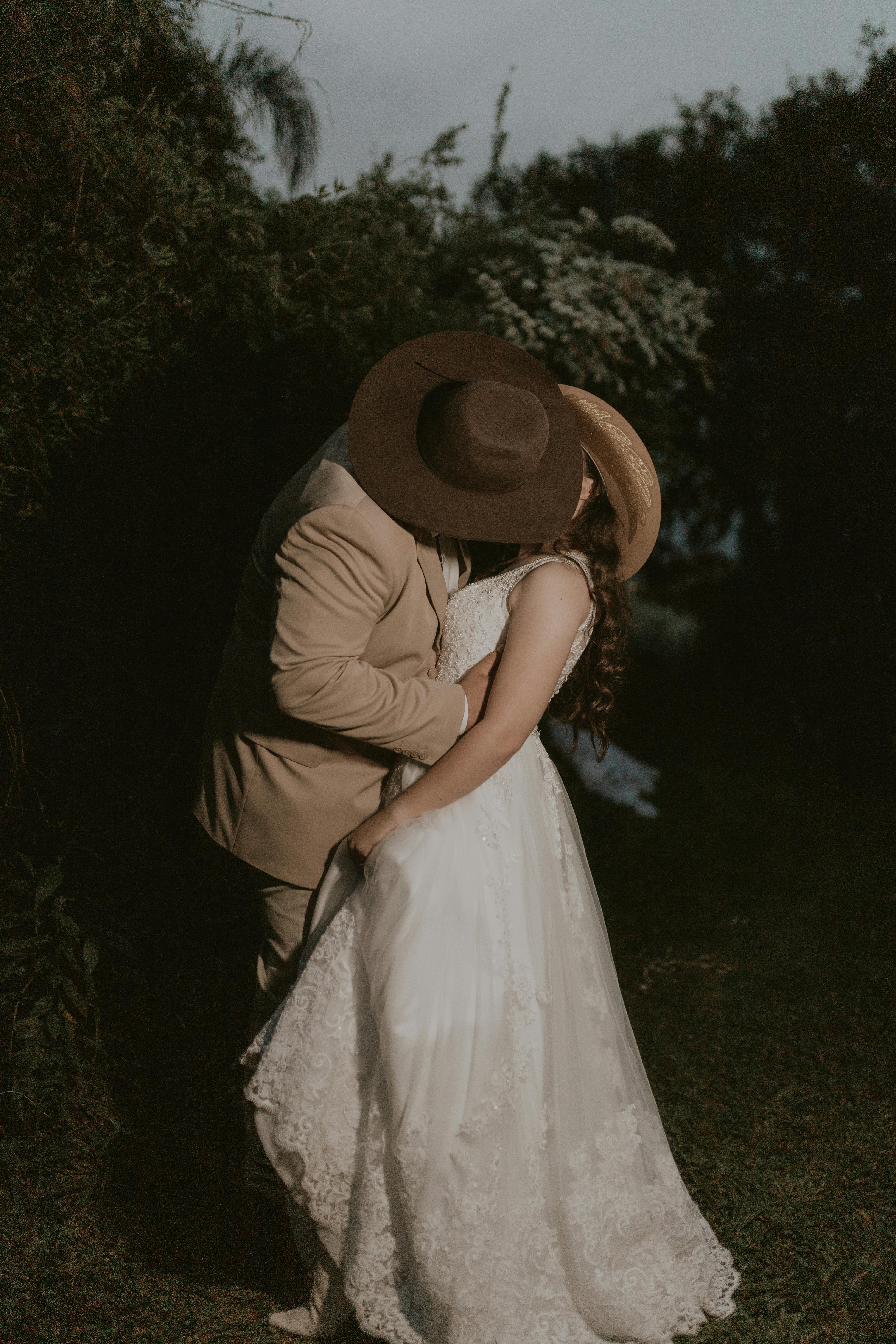 A couple in wedding attire shares a romantic kiss outdoors during twilight.