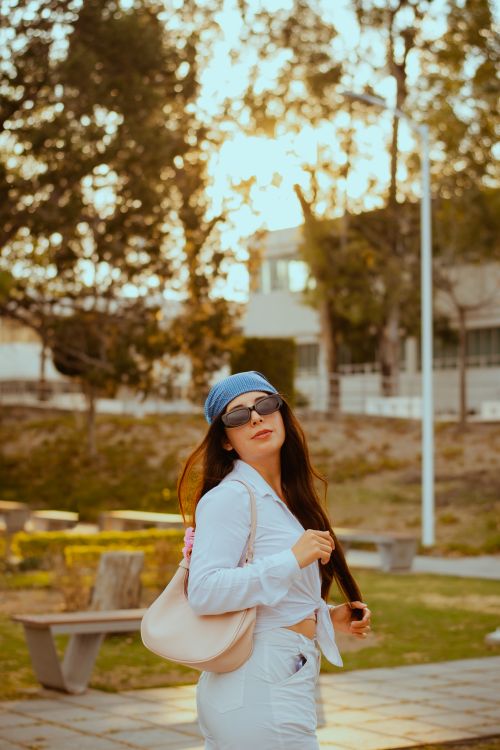 Free A fashionable woman wearing sunglasses poses confidently in a sunlit park setting. Stock Photo
