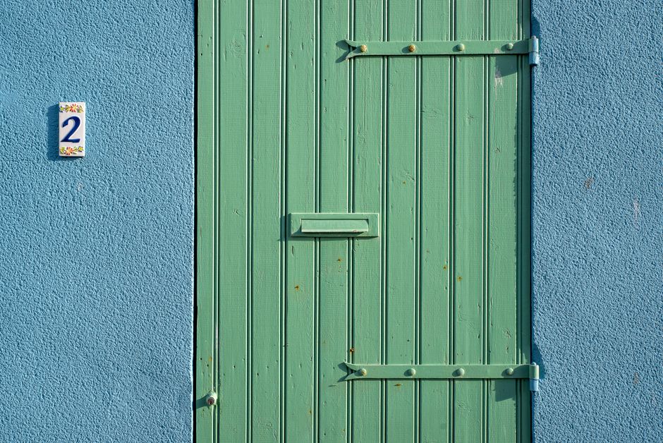 A green wooden door with metal hinges against a blue textured wall featuring a street number.