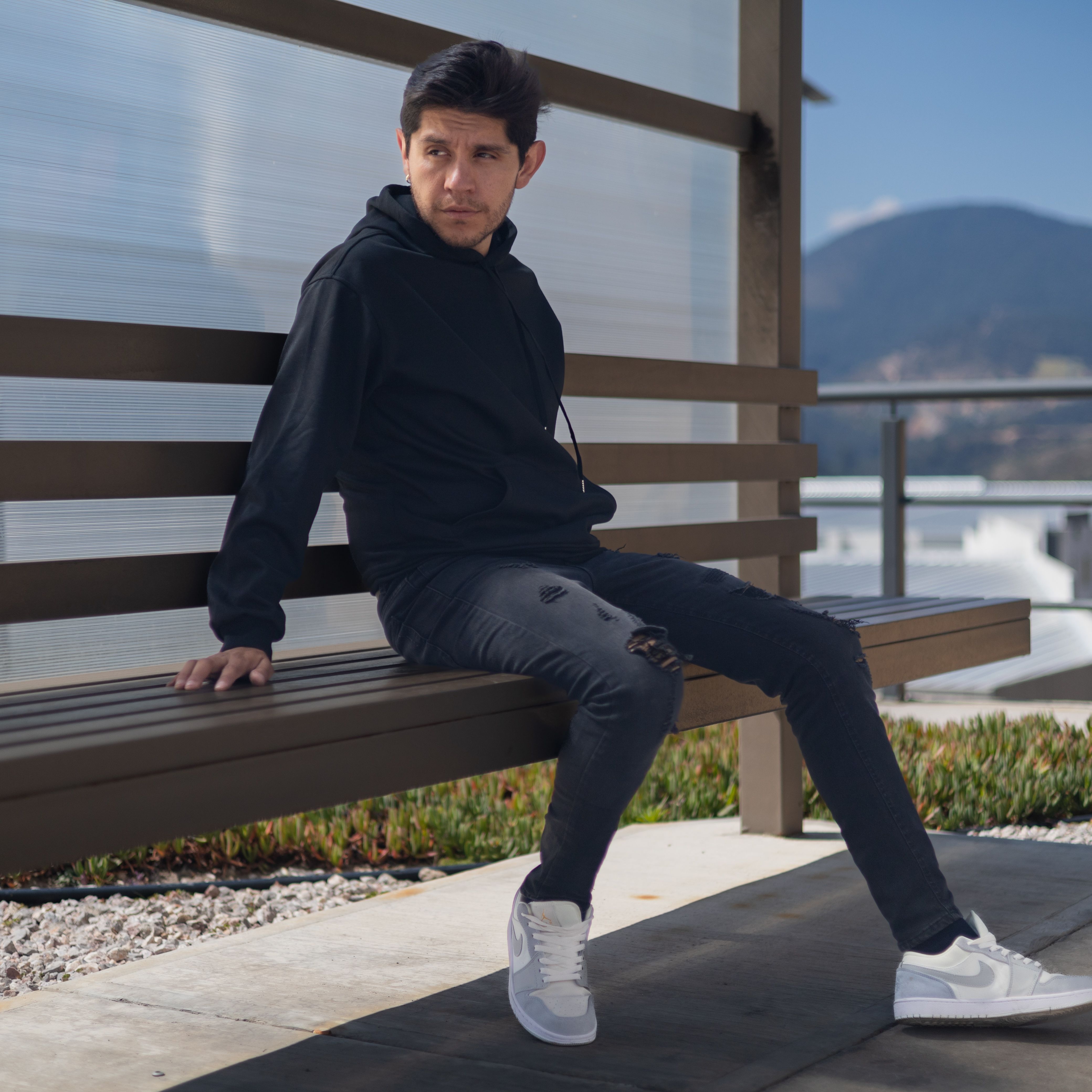 Stylish male in a black hoodie sits on a bench outdoors. Mountain view in background.